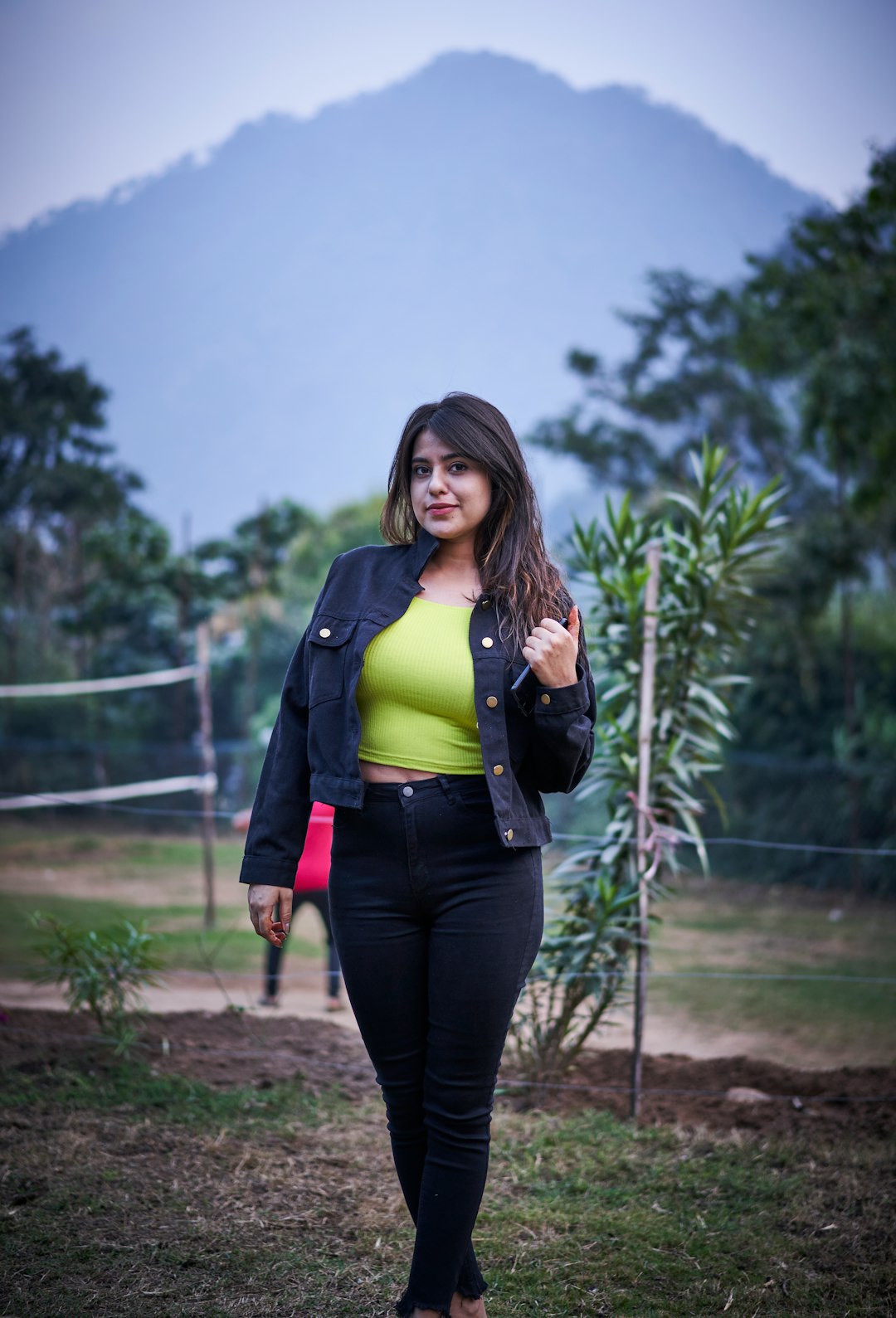 smiling woman wearing green top and black denim jacket standing on green field