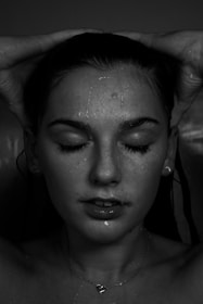 A close-up portrait of a person with water droplets on their face. Their eyes are closed, and hands are gently placed on their head. The lighting is dramatic with a focus on the facial features and water, creating a serene expression.
