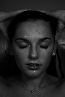 A close-up portrait of a person with water droplets on their face. Their eyes are closed, and hands are gently placed on their head. The lighting is dramatic with a focus on the facial features and water, creating a serene expression.