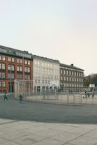 A public square with children playing near a fenced area. The background features three distinct, multistory buildings with traditional architecture in red, white, and brown colors. The scene reflects a calm, urban setting with a cloudy sky.