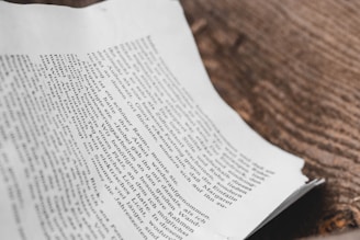 A close-up view of a printed page with a portion of text in a serif font, lying on a wooden surface. The text appears to be in a language using Latin script, and the page is slightly curled at the edges.