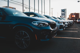 A row of parked luxury cars, primarily in dark and silver colors, is aligned in a parking lot during the evening. The setting features several identical car models, and dealership signage is visible in the background, indicating a commercial car lot.