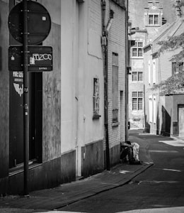A narrow alleyway between buildings with a homeless person sitting against a wall. The buildings are old with visible wear and graffiti on the walls. The street sign is defaced with graffiti. The scene is captured in black and white, giving it a somber and gritty appearance.