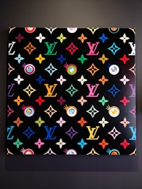 A vibrant, patterned artwork featuring repeated motifs and symbols in multiple colors against a black background. The pattern includes various geometric shapes and floral designs, with a focus on a recognizable monogram repeated in different hues across the canvas.