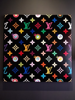 A vibrant, patterned artwork featuring repeated motifs and symbols in multiple colors against a black background. The pattern includes various geometric shapes and floral designs, with a focus on a recognizable monogram repeated in different hues across the canvas.