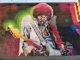 A vibrantly colored mural featuring a musician with an afro hairstyle playing an electric guitar. The artwork showcases intricate details such as a patterned strap and a collared shirt. The wall includes green and blue accents with a textured background.