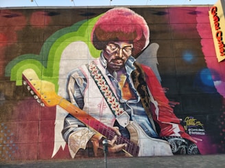 A vibrantly colored mural featuring a musician with an afro hairstyle playing an electric guitar. The artwork showcases intricate details such as a patterned strap and a collared shirt. The wall includes green and blue accents with a textured background.