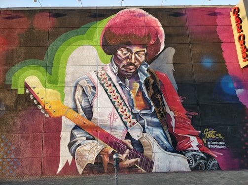 A vibrantly colored mural featuring a musician with an afro hairstyle playing an electric guitar. The artwork showcases intricate details such as a patterned strap and a collared shirt. The wall includes green and blue accents with a textured background.