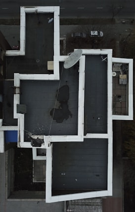 An aerial view of a rooftop with a complex geometric layout. The roof features a white border around a darker central surface. A large satellite dish is positioned near the center, with some visible water pooling on part of the roof. The surrounding area includes a street with a couple of parked cars and some sparse vegetation.