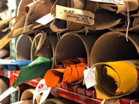 Rolled-up pieces of leather in various colors are stored on large cylindrical shelves. Each piece has a small white tag with handwritten details attached, indicating specific information like texture or type. The leather colors include shades of brown, green, blue, orange, and yellow.