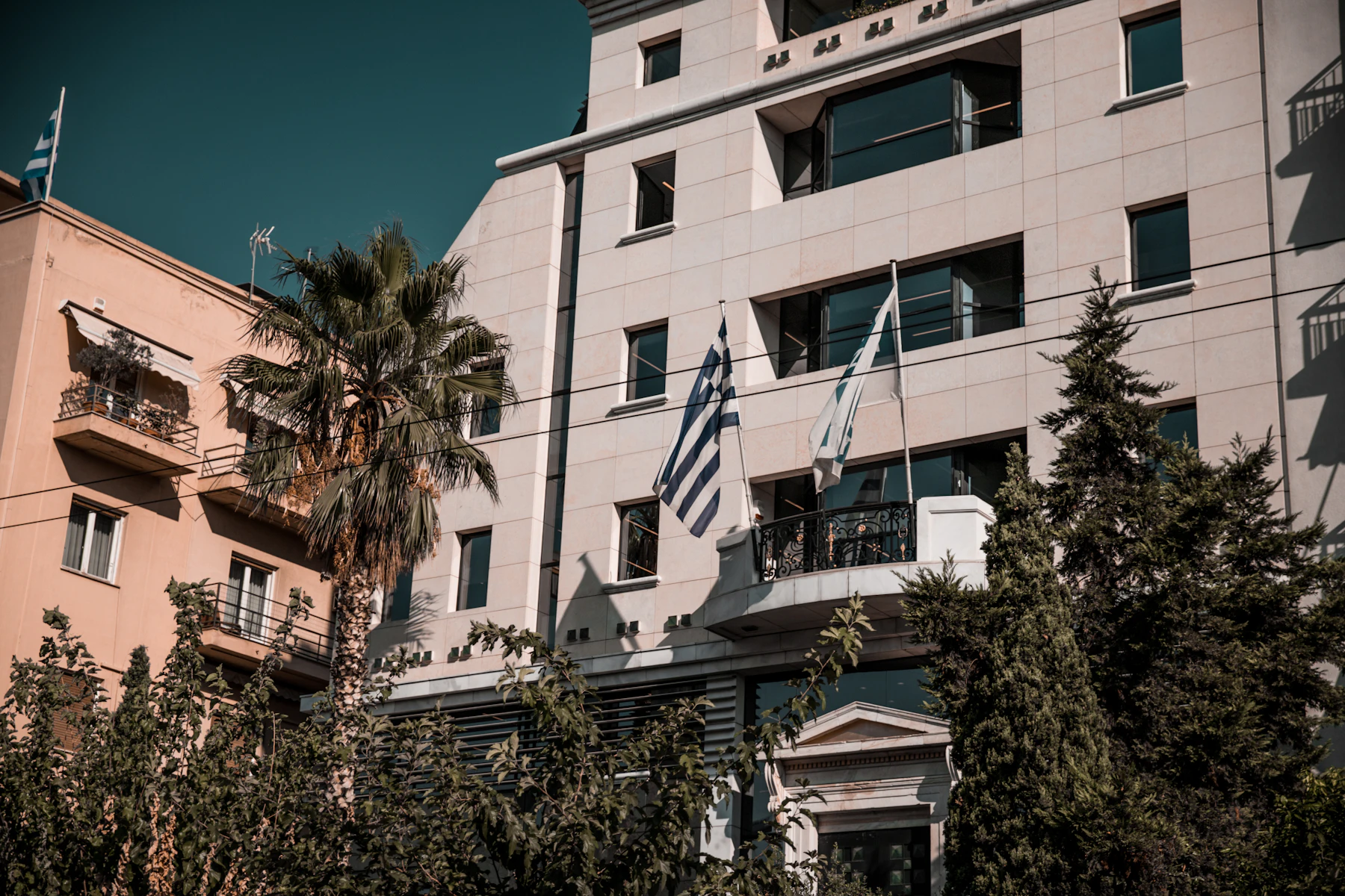 An Athens building flying the Greek and European flags, palm and shuttered balconies against a hard blue sky.
