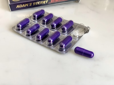 A blister pack containing several purple capsules sits on a white surface. The pack has one capsule missing, which is placed next to it. In the background, there is a box with the label 'Adam's Secret' and a golden badge, partially visible.