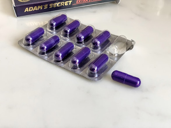 A blister pack containing several purple capsules sits on a white surface. The pack has one capsule missing, which is placed next to it. In the background, there is a box with the label 'Adam's Secret' and a golden badge, partially visible.