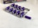 A blister pack containing several purple capsules sits on a white surface. The pack has one capsule missing, which is placed next to it. In the background, there is a box with the label 'Adam's Secret' and a golden badge, partially visible.