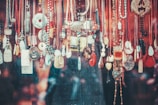 A variety of necklaces and pendants hang closely together against a textured background. The jewelry pieces feature diverse designs, materials, and colors, including beads, stones, and intricately carved pendants. Several cords are visible, most prominently in red, black, and brown shades. The overall arrangement appears artistic and eclectic.