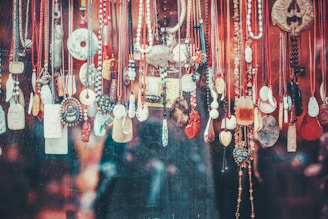 A variety of necklaces and pendants hang closely together against a textured background. The jewelry pieces feature diverse designs, materials, and colors, including beads, stones, and intricately carved pendants. Several cords are visible, most prominently in red, black, and brown shades. The overall arrangement appears artistic and eclectic.