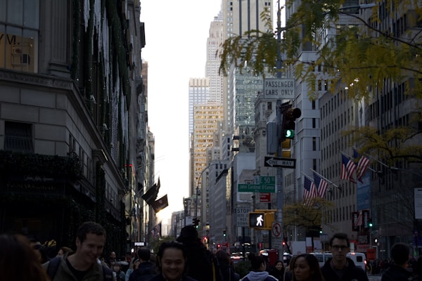 A bustling city street with tall skyscrapers framing the scene, numerous people walking along the sidewalk, and several American flags attached to buildings. Traffic signals and street signs are visible, with leaves from a tree adding a touch of nature to the urban environment.