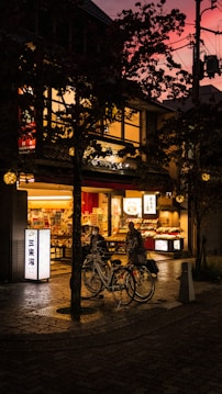 A cozy street scene at dusk, showcasing a warmly lit storefront with bicycles parked in front. The evening sky has a vibrant pink hue, contrasting with the darker silhouettes of surrounding trees. The scene is alive with subtle reflections on the wet pavement and muted street lights.