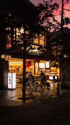 A cozy street scene at dusk, showcasing a warmly lit storefront with bicycles parked in front. The evening sky has a vibrant pink hue, contrasting with the darker silhouettes of surrounding trees. The scene is alive with subtle reflections on the wet pavement and muted street lights.