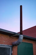A metal chimney extends vertically from the side of a brick building against a twilight sky. The structure has a reddish-brown tone with a curved segment. The wall also features a green door and a mesh-covered window.