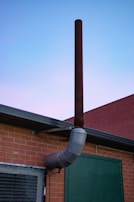 A metal chimney extends vertically from the side of a brick building against a twilight sky. The structure has a reddish-brown tone with a curved segment. The wall also features a green door and a mesh-covered window.