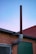 A metal chimney extends vertically from the side of a brick building against a twilight sky. The structure has a reddish-brown tone with a curved segment. The wall also features a green door and a mesh-covered window.