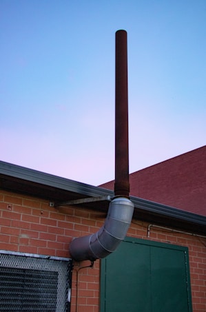 A metal chimney extends vertically from the side of a brick building against a twilight sky. The structure has a reddish-brown tone with a curved segment. The wall also features a green door and a mesh-covered window.