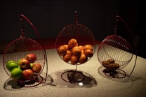 Three spherical wire baskets with metal stands are arranged in a row on a speckled countertop. The left basket contains a mix of green and red apples, the center one holds several oranges, and the right basket has a few brown kiwis. Dim lighting creates a warm and cozy atmosphere with soft shadows cast around the objects.