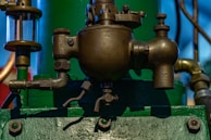 A close-up view of a metallic industrial component, possibly part of a machine, mounted on a green base. The component has multiple valves, a pipe extending from the left side, and an intricate assembly with various bolts and connectors. The background features blurred objects in different colors, including blue and red.