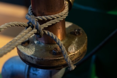 A metallic pipe is secured with a thick rope wrapped around its base. The rope appears frayed and well-worn, suggesting frequent use. The pipe and its fittings are shiny with a warm reflection from a nearby light source, highlighting textures on the metal surface.