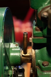 A close-up of a metallic mechanical component featuring green painted parts, a brass or bronze fitting, and a threaded pipe extending vertically. The focus is on the industrial equipment, showing the textures and details of the metal surfaces.