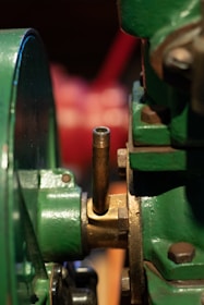 A close-up of a metallic mechanical component featuring green painted parts, a brass or bronze fitting, and a threaded pipe extending vertically. The focus is on the industrial equipment, showing the textures and details of the metal surfaces.