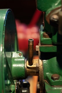 A close-up of a metallic mechanical component featuring green painted parts, a brass or bronze fitting, and a threaded pipe extending vertically. The focus is on the industrial equipment, showing the textures and details of the metal surfaces.