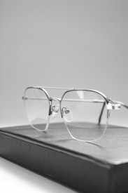 A pair of rimless eyeglasses rests on a closed book. The glasses have a minimalist design with clear lenses and metallic frames. The book appears to have a dark cover, contrasting with the light, soft-focus background.