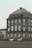 A large, historic building with a brown, multi-story facade and white-trimmed windows is in the background. In front of it, a person is riding a gray horse, and another white horse stands nearby in an outdoor enclosure or riding arena.