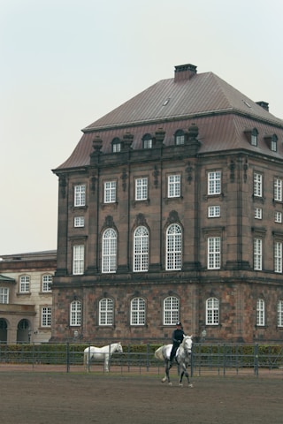 A large, historic building with a brown, multi-story facade and white-trimmed windows is in the background. In front of it, a person is riding a gray horse, and another white horse stands nearby in an outdoor enclosure or riding arena.
