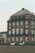 A large, historic building with a brown, multi-story facade and white-trimmed windows is in the background. In front of it, a person is riding a gray horse, and another white horse stands nearby in an outdoor enclosure or riding arena.
