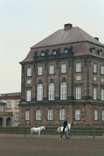 A large, historic building with a brown, multi-story facade and white-trimmed windows is in the background. In front of it, a person is riding a gray horse, and another white horse stands nearby in an outdoor enclosure or riding arena.