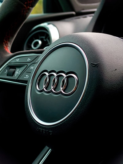 A close-up view of a car steering wheel featuring the Audi logo. Visible are the circular air vents and part of the dashboard, with a sleek, modern design.