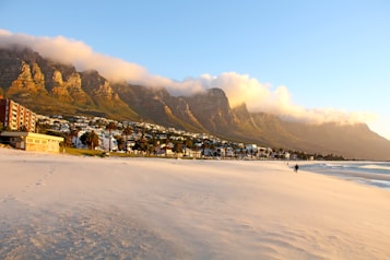 A serene beach with white sand stretches along the foreground, with footprints trailing towards the shoreline. The beach is lined with buildings and palm trees. Majestic mountains with peaks partially covered by clouds dominate the background, basking in the warm, golden light of either sunrise or sunset. A few people are walking along the beach, creating a tranquil and picturesque scene.