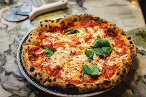 A freshly baked pizza with a thick, golden-brown crust and topped with melted mozzarella cheese, tomato sauce, and fresh basil leaves. The pizza is presented on a decorative plate placed on a marble surface. A pizza cutter with a white handle lies nearby.