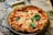 A freshly baked pizza with a thick, golden-brown crust and topped with melted mozzarella cheese, tomato sauce, and fresh basil leaves. The pizza is presented on a decorative plate placed on a marble surface. A pizza cutter with a white handle lies nearby.