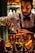 A bartender is carefully pouring liquid from a small copper jigger into a shaker. The individual is dressed in a white shirt with a bow tie and dark suspenders. In the foreground, several glasses contain garnishes such as lemon and lime slices. The background features a softly lit bar with bottles arranged on shelves.
