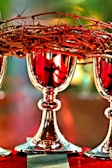 A crown of thorns is placed atop silver chalices, with reflections showcasing a cross. The lighting creates a dramatic and sacred atmosphere, emphasizing the intricate details of the thorns and the polished shine of the chalices.
