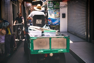 A person wearing a straw hat is driving a small three-wheeled vehicle loaded with white buckets and miscellaneous items. The vehicle is moving through a narrow alley lined with closed shop doors and signs. Another person pushes a bicycle alongside, and two individuals, including a child wearing a hat, are seen on the left side. The alley is shaded but bright with sunlight filtering through.