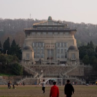 A large, traditional-style building with grand architectural features and a prominent sign on its facade. It is surrounded by trees and set against a backdrop of a hill. People are visible in the foreground, engaging in leisure activities on an open, grassy area.
