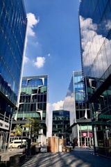A cityscape featuring modern glass buildings with sharp geometric designs reflecting the clear blue sky and clouds. The scene is vibrant with people walking along the street, surrounded by small trees and outdoor seating. Commercial spaces with colorful signage are visible, contributing to a lively urban atmosphere.