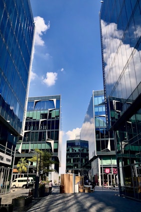 A cityscape featuring modern glass buildings with sharp geometric designs reflecting the clear blue sky and clouds. The scene is vibrant with people walking along the street, surrounded by small trees and outdoor seating. Commercial spaces with colorful signage are visible, contributing to a lively urban atmosphere.