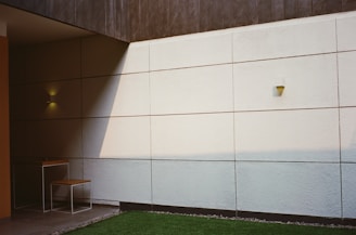 A minimalist patio area featuring a concrete wall with square panels, partially illuminated by soft, natural light casting angular shadows. A small, modern table and chair set with a thin metal frame and wooden surfaces is placed in the corner. The ground is covered with a patch of green grass bordered by small rocks.