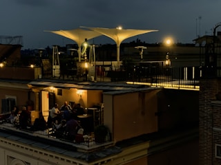 A rooftop terrace is dimly lit with warm, ambient lights during the evening. People are gathered around tables, engaged in conversation and enjoying a relaxed atmosphere. The terrace features modern outdoor furniture and large, white, triangular shade structures. The sky is dark, suggesting nighttime.